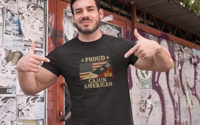 Man wearing a 'Proud Cajun American' t-shirt in front of a wall with posters.